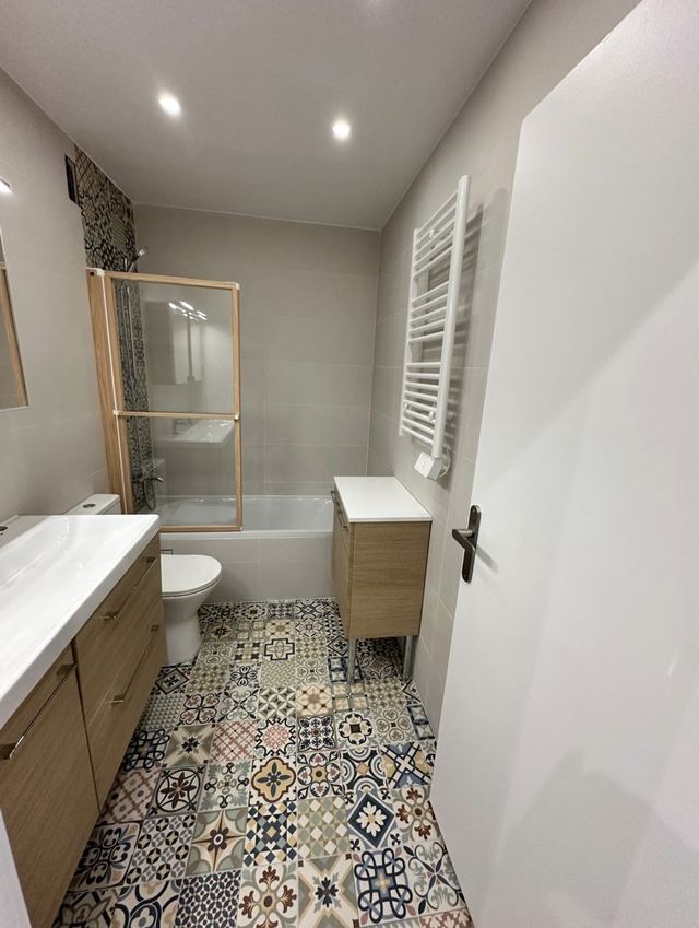 Salle de bain avec baignoire et pare-bain, sol en carreaux de ciment à motifs, meubles bois et sèche-serviettes